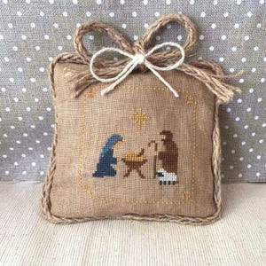 May include: A square, beige fabric ornament with a cross-stitch nativity scene. The scene depicts Mary, Joseph, and baby Jesus in a manger, with a star above. The ornament has a braided rope border and a jute bow at the top.