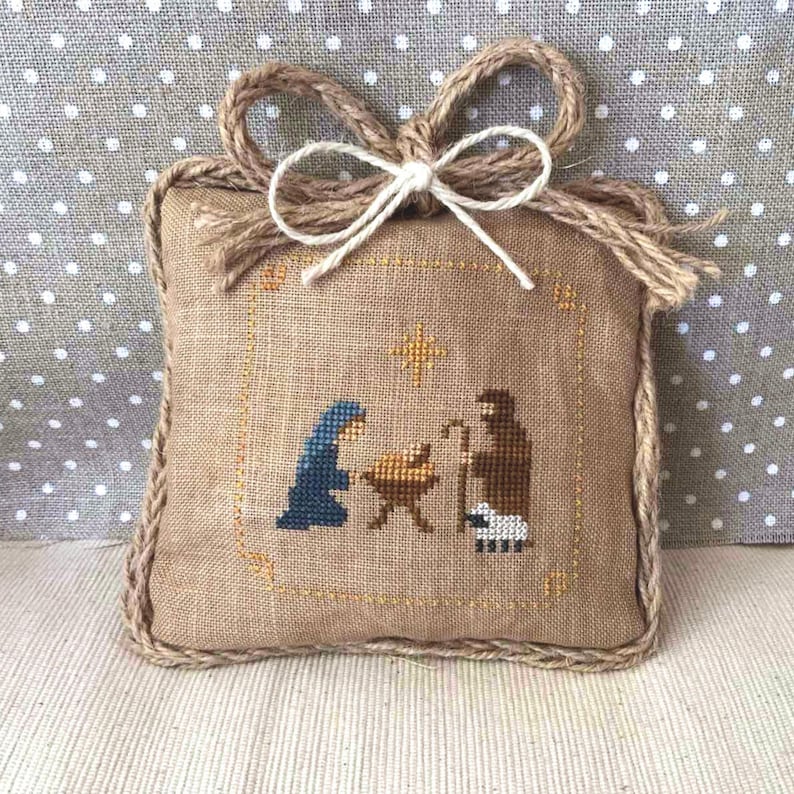 May include: A square, beige fabric ornament with a cross-stitch nativity scene. The scene depicts Mary, Joseph, and baby Jesus in a manger, with a star above. The ornament has a braided rope border and a jute bow at the top.