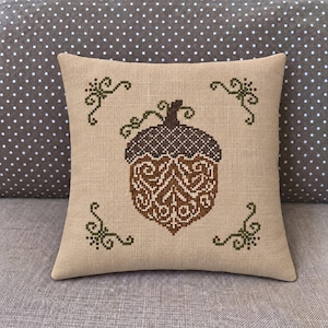 May include: A square throw pillow made of light tan linen fabric featuring a cross-stitched acorn design in the center. The acorn is rendered in shades of brown with dark green decorative scrollwork accents in each of the four corners.