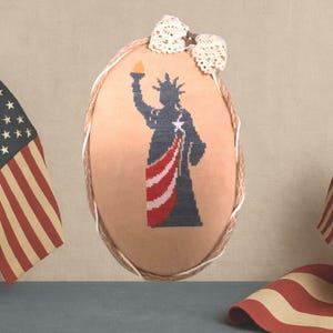 Peut inclure: Décoration murale ovale avec une broderie au point de croix de la Statue de la Liberté. Le motif utilise les couleurs rouge, blanc et bleu. La décoration a une bordure en corde et un nœud en dentelle crème en haut.