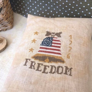 Può includere: Un cuscino beige con un disegno a punto croce che presenta una campana con una bandiera americana, stelle e il testo "1776 FREEDOM".
