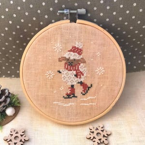 May include: A cross-stitch embroidery hoop featuring a white snowman wearing a red hat and scarf, ice skating on a white background with snowflakes.