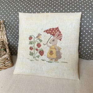 May include: A square, cream-colored pillow with a cross-stitched design. The design features a bunny holding a red umbrella with white polka dots, a bird, and a strawberry plant. The bunny wears a yellow apron.