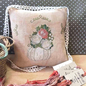 May include: A white pumpkin with a pink rose and green leaves embroidered on a beige pillow with white lace trim. The pillow is on a brown and white polka dot fabric.