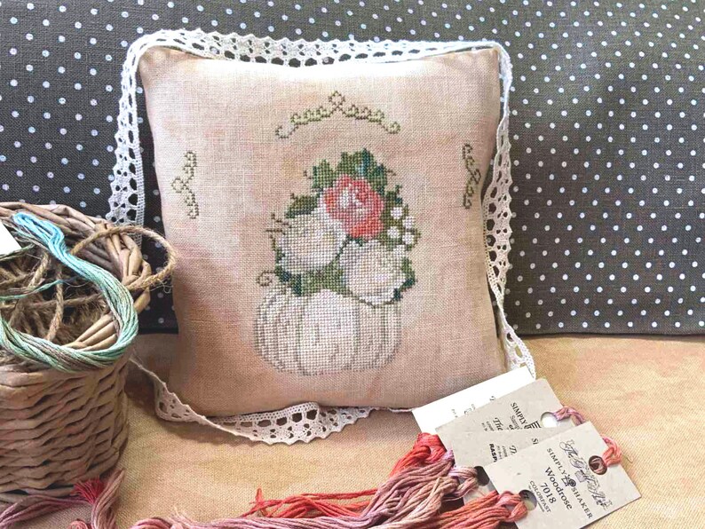 May include: A white pumpkin with a pink rose and green leaves embroidered on a beige pillow with white lace trim. The pillow is on a brown and white polka dot fabric.