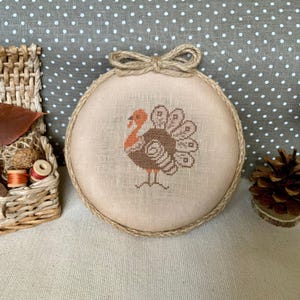Puede incluir: Un bastidor de bordado de punto de cruz redondo y beige con un diseño de pavo marrón y naranja. El bastidor está adornado con hilo de bramante y tiene un lazo de hilo de bramante en la parte superior. A la izquierda hay una pequeña cesta con útiles de costura.