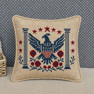 May include: A square, beige pillow with a cross-stitched design featuring a blue eagle, red and blue shield, red stars, and floral accents. The pillow has a rope-like border.