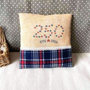 May include: Square decorative pillow with a tan linen top, embroidered with red and blue stars forming the number "20". The bottom half features a blue, red, and white plaid pattern, with a white lace trim separating the two sections. The text "1776 2026" is embroidered below the number.