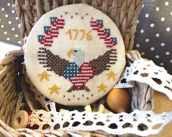 May include: A white fabric circle with a cross-stitch design of an American bald eagle with a red, white, and blue heart, surrounded by small American flags and the year 1776 in yellow thread.