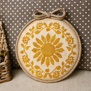 May include: A circular embroidery hoop containing a cross-stitch design of a large central sunflower surrounded by a floral wreath. The stitching is done in golden yellow thread on a cream-colored fabric. A jute rope trim edges the hoop.