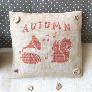 May include: A beige pillow with the word "AUTUMN" embroidered in brown. The pillow features a red squirrel and a red gramophone with musical notes embroidered on it.