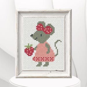 Może przedstawiać: Haft krzyżykowy przedstawiający szarą mysz w różowej sukience i czerwonej kokardzie, trzymającą czerwoną truskawkę. Oprawiony obraz ma białą, postarzaną drewnianą ramę. Grafika jest prezentowana na białej powierzchni.