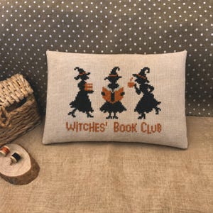 May include: A rectangular beige linen pillow featuring a cross-stitch design of three black silhouettes of witches wearing pointed hats. The witches are holding an orange book and a mug. The text Witches' Book Club is stitched in orange.