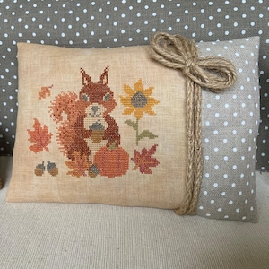 May include: A cross-stitched pillow featuring a brown squirrel holding an acorn, a pumpkin, and a sunflower. The design includes fall leaves in shades of orange and red. The pillow is tied with twine and has a polka dot backing.
