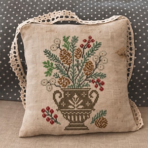 May include: A square beige linen cushion featuring a cross-stitched design of a dark brown urn filled with pinecones, green pine needles, and red and white berries. The cushion is edged with a decorative cream-coloured crocheted lace trim.
