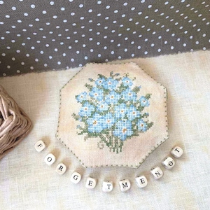 May include: A white fabric hexagon-shaped needlepoint with a cross-stitch design of a bouquet of blue forget-me-not flowers. The needlepoint is surrounded by white wooden letter tiles spelling out "Forget Me Not".