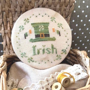 May include: A round, white, cross-stitched piece of fabric with a green leprechaun hat and the word "Irish" in green thread. The fabric is in a wicker basket with a white cloth and yellow thread.