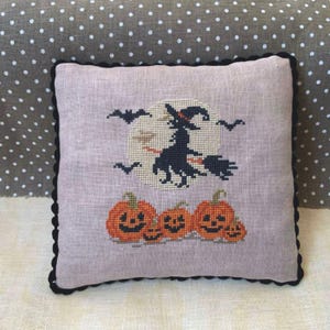 Może przedstawiać: Dekoracyjna poduszka z haftowanym wzorem czarownicy latającej na miotle przed pełnią księżyca. Wzór zawiera również cztery pomarańczowe dynie z twarzami dyni.