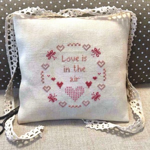 May include: A white pillow with a red embroidered design of hearts and the text "Love is in the air". The pillow is trimmed with white lace and is sitting on a brown fabric with white polka dots.