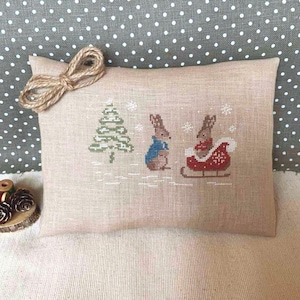 Peut inclure: Un coussin rectangulaire beige avec une scène d'hiver brodée au point de croix. Deux lapins sont représentés, l'un avec un manteau bleu et l'autre dans un traîneau rouge. Un sapin vert et des flocons de neige complètent le motif.