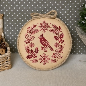 May include: A circular embroidery hoop featuring a cross-stitch design on beige fabric. The pattern displays a dark red cardinal bird centered within a wreath of holly leaves, berries, and snowflakes. A jute twine bow is tied at the top of the hoop.