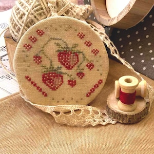 May include: A close-up of a hand-stitched embroidery hoop with a design of red strawberries on a beige background. The hoop is surrounded by a white lace trim and a spool of red thread.