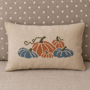 May include: A rectangular beige linen pillow featuring a cross-stitch embroidery design of five pumpkins in shades of orange and blue. The pumpkins are arranged in a cluster with green stems and vines, centered on the front of the pillow cover.
