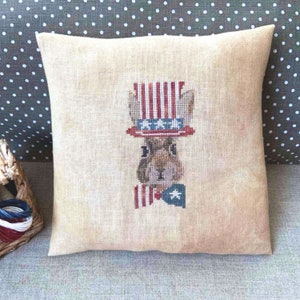 May include: A beige linen pillow with a cross-stitch design of a brown bunny wearing a red, white, and blue hat and bow tie. The hat has red and white stripes and white stars. The bow tie has red and white stripes and a blue star.