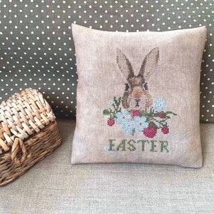 K&ouml;nnte beinhalten: Ein beigefarbenes Kissen mit einem Kreuzstich-Design eines braunen Kaninchens mit wei&szlig;en Blumen und roten Erdbeeren. Das Kissen ist mit dem Wort "Easter" bestickt.