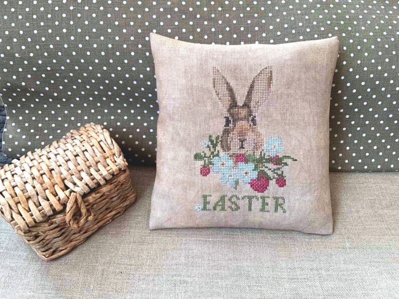 K&ouml;nnte beinhalten: Ein beigefarbenes Kissen mit einem Kreuzstich-Design eines braunen Kaninchens mit wei&szlig;en Blumen und roten Erdbeeren. Das Kissen ist mit dem Wort "Easter" bestickt.