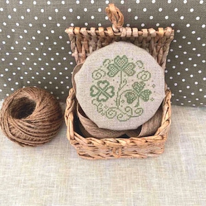 May include: A round, hand-stitched piece of fabric with a green shamrock design in a woven basket. The basket is on a brown and white polka dot fabric.