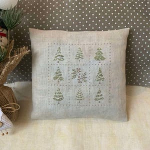 Peut inclure: Un coussin en lin beige avec un motif de point de croix de neuf arbres verts et un flocon de neige au centre. Le coussin est sur un tissu à pois brun et blanc.