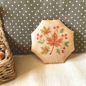 Puede incluir: Un objeto decorativo octogonal beige bordado a punto de cruz con hojas de otoño en tonos naranja, amarillo y verde, con pequeñas bayas rojas. El objeto está sobre una superficie blanca, con una cesta en el fondo.