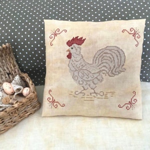 Puede incluir: Un gallo blanco con cresta y barbillas rojas está bordado en una almohada beige con un fondo de lino marrón. El gallo está rodeado de cuatro remolinos rojos.