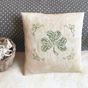 May include: A beige linen pillow with a green embroidered shamrock design. The shamrock is surrounded by four green swirls.
