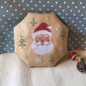 Może przedstawiać: Ręcznie robiona, sześciokątna ozdoba świąteczna z haftowanym krzyżykiem wzorem Świętego Mikołaja. Ozdoba ma beżowe tło z czerwonym kapeluszem, białą brodą i zielonymi drzewami. Na ozdobie wyhaftowano również płatki śniegu.