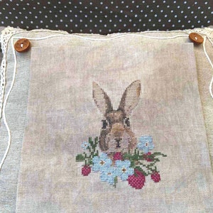 K&ouml;nnte beinhalten: Ein beigefarbenes Leinenkissen mit einer Kreuzstich-Stickerei eines braunen Kaninchens mit blauen Blumen und roten Beeren.