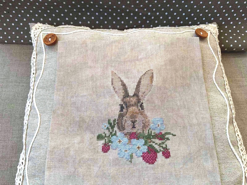 K&ouml;nnte beinhalten: Ein beigefarbenes Leinenkissen mit einer Kreuzstich-Stickerei eines braunen Kaninchens mit blauen Blumen und roten Beeren.