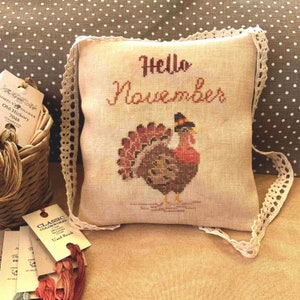 Op de afbeelding: Een beige kussen met een witte kantversiering heeft een kruissteekontwerp van een kalkoen met de tekst "Hello November".