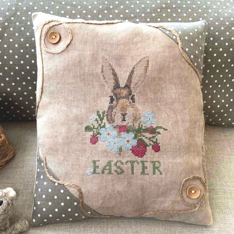 K&ouml;nnte beinhalten: Ein beigefarbenes Kissen mit einem braunen Hasen, der darauf gestickt ist. Der Hase ist von Blumen und Erdbeeren umgeben. Das Kissen hat einen braunen Jutesaum und zwei braune Kn&ouml;pfe. Das Wort "Easter" ist unter dem Hasen gestickt.