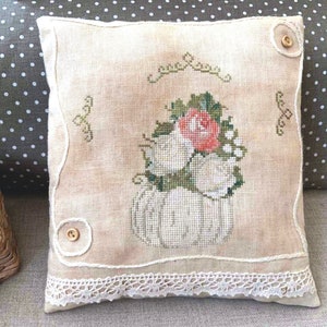 May include: A white linen pillow with a cross-stitch design of a white pumpkin with a bouquet of white roses and a single pink rose. The pillow has a lace trim and two brown buttons.