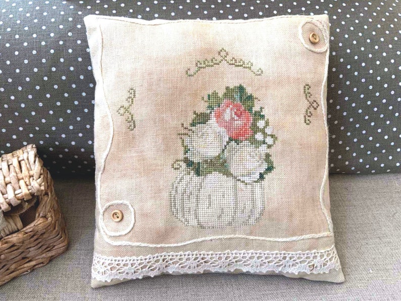 May include: A white linen pillow with a cross-stitch design of a white pumpkin with a bouquet of white roses and a single pink rose. The pillow has a lace trim and two brown buttons.