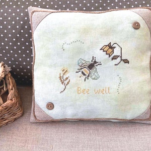 May include: A light green pillow with a brown border and two brown buttons. The pillow has an embroidered bee, flowers, and the text "Bee well".
