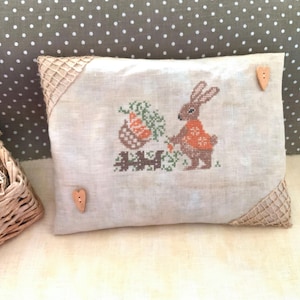 Puede incluir: Una almohada beige con ribete de arpillera marrón y dos botones de madera en forma de corazón. La almohada presenta un diseño bordado de un conejo que lleva una cesta de zanahorias.