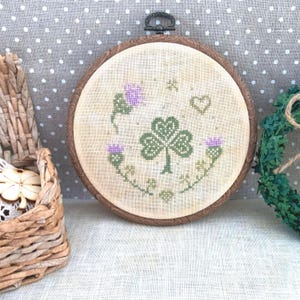 May include: A circular embroidery hoop with a white fabric background featuring a green shamrock with three leaves, surrounded by three purple thistles and small green shamrocks. The hoop is made of wood with a brown stain.