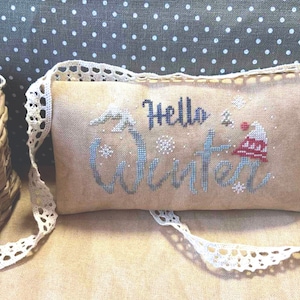 May include: A rectangular, beige cross-stitch pillow with the words "Hello Winter" in blue and white thread. The pillow features snowflakes and a red and white hat design. It has a cream-colored lace strap.