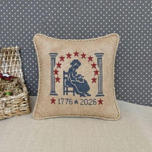 Puede incluir: Un cojín cuadrado beige con un diseño de punto de cruz. El diseño presenta una silueta azul de una mujer sentada, rodeada de estrellas y pilares rojos. Los números "1776" y "2026" están debajo. El cojín tiene un ribete de cuerda.