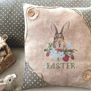 K&ouml;nnte beinhalten: Ein beiges Kissen mit einem braunen Hasen, der darauf gestickt ist. Der Hase ist von Blumen und Erdbeeren umgeben. Das Kissen hat einen braunen Stoffrand und das Wort "Easter" ist unter dem Hasen gestickt.