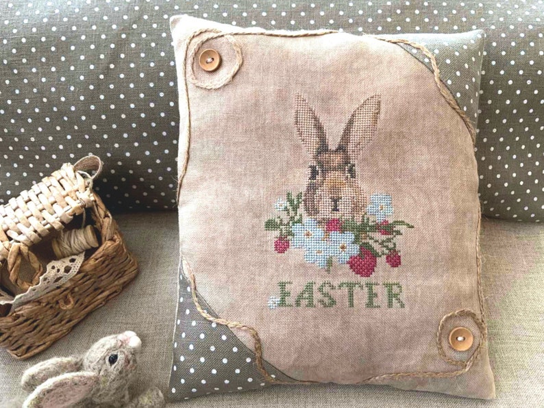 K&ouml;nnte beinhalten: Ein beiges Kissen mit einem braunen Hasen, der darauf gestickt ist. Der Hase ist von Blumen und Erdbeeren umgeben. Das Kissen hat einen braunen Stoffrand und das Wort "Easter" ist unter dem Hasen gestickt.