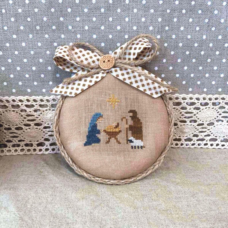 May include: A round, cross-stitched Christmas ornament featuring a nativity scene with Mary, Joseph, and baby Jesus. The ornament has a tan background, a brown rope border, and a decorative bow with a button. The bow has a brown and white checkered pattern.